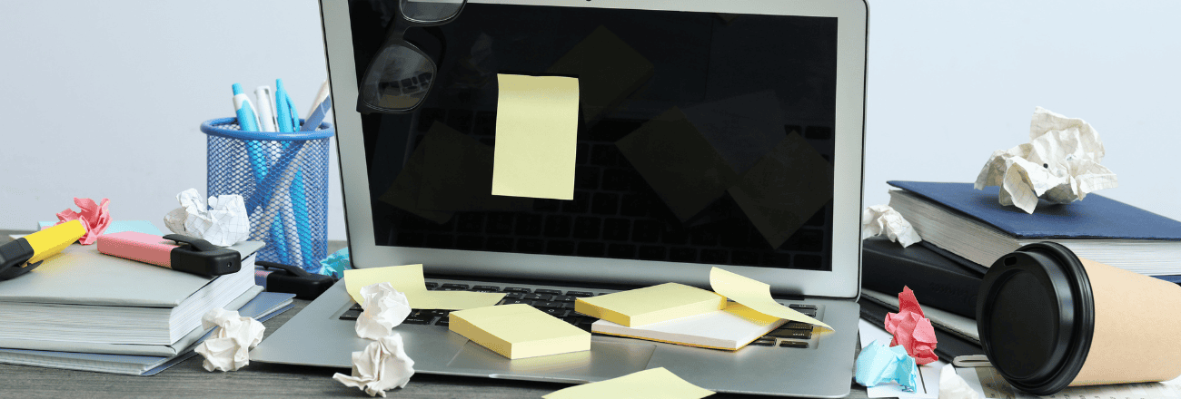 Messy desk with sticky notes on laptop, crumpled papers, and a coffee cup